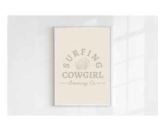 May include: A framed print with a beige background and a gray logo that says "SURFING COWGIRL Montauk Brewing Co. New York". The logo features a stylized seashell.