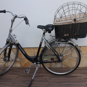 Puede incluir: Una bicicleta Gazelle plateada y negra con una gran cesta de mimbre y una jaula de alambre en la parte trasera. La bicicleta tiene un asiento, guardabarros y manillar negros. La bicicleta est&aacute; aparcada sobre una superficie de madera.
