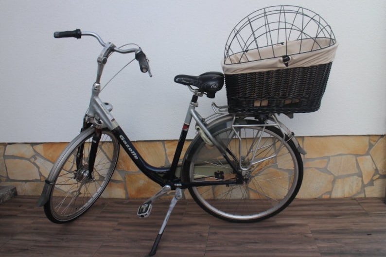 Puede incluir: Una bicicleta Gazelle plateada y negra con una gran cesta de mimbre y una jaula de alambre en la parte trasera. La bicicleta tiene un asiento, guardabarros y manillar negros. La bicicleta est&aacute; aparcada sobre una superficie de madera.