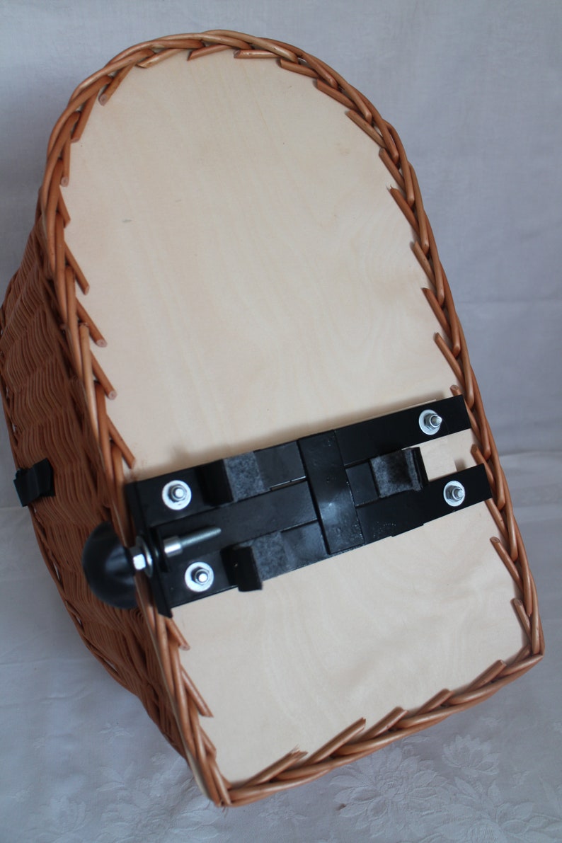 Puede incluir: Una cesta de mimbre para bicicleta con una base de madera de color claro. La cesta tiene un exterior tejido marr&oacute;n y un soporte de montaje de metal negro con pernos. La cesta est&aacute; dise&ntilde;ada para sujetarse a una bicicleta.