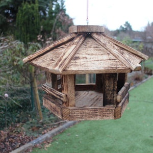 May include: A wooden birdhouse with a shingled roof and a small opening for birds to enter. The birdhouse is brown and has a weathered look. It is hanging from a string and is in front of a green lawn.