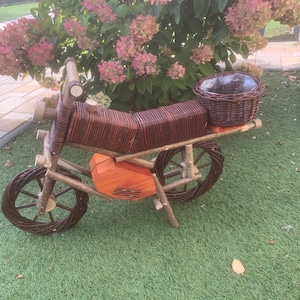 Op de afbeelding: Een decoratieve houten motorfiets bloempot met een bruine geweven zitting en mand. De wielen en het frame zijn gemaakt van natuurlijk hout, en de carrosserie heeft een oranje houten kist. Ontworpen voor bloemen of kleine planten.