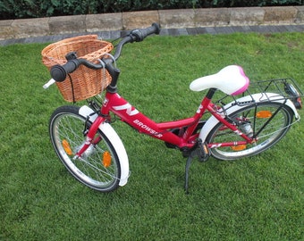 Puede incluir: Una bicicleta roja con una cesta de mimbre blanca, guardabarros blancos y un sillín blanco con ribete rosa. La bicicleta tiene un portaequipajes negro en la parte trasera y las palabras "Browstar" en el cuadro.