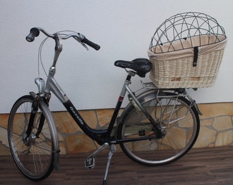 Puede incluir: Una bicicleta plateada con un cuadro negro y una cesta de mimbre con una jaula de alambre negro en la parte trasera. La bicicleta tiene un sillín negro y una pata de cabra.