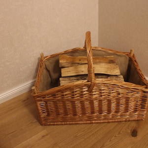Könnte beinhalten: Ein hellbrauner Weidenkorb mit Griff, gefüllt mit Brennholz. Der Korb ist mit einem Jutematerial ausgekleidet. Der Korb steht auf einem Holzboden.