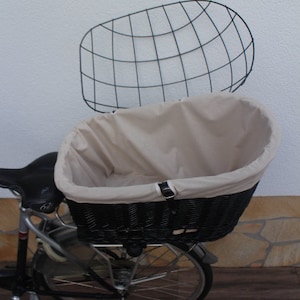 Puede incluir: Una cesta de bicicleta de mimbre negro con un forro de tela beige y una rejilla de metal negro a juego. La cesta est&aacute; montada en una bicicleta, ofreciendo una soluci&oacute;n de almacenamiento elegante y pr&aacute;ctica para el ciclismo.