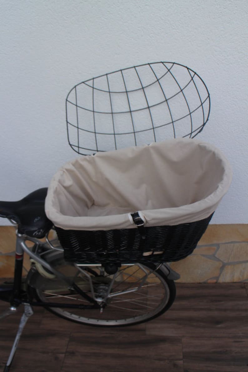 Puede incluir: Una cesta de bicicleta de mimbre negro con un forro de tela beige y una rejilla de metal negro a juego. La cesta est&aacute; montada en una bicicleta, ofreciendo una soluci&oacute;n de almacenamiento elegante y pr&aacute;ctica para el ciclismo.