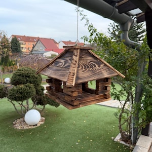 May include: A hanging wooden birdhouse with a dark, charred finish. The birdhouse has a peaked roof and open sides, suspended by a thin wire. The background shows a green lawn, trees, and houses.