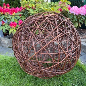 Könnte beinhalten: Eine große, kugelförmige Gartendekoration aus verflochtenen braunen Ästen. Die kugelartige Skulptur liegt auf grünem Gras, mit leuchtend rosa Blüten und Blättern im Hintergrund. Dieses natürliche Element verleiht Außenbereichen einen rustikalen Touch.