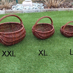 Op de afbeelding: Drie rieten manden in verschillende maten, gelabeld met XXL, XL en L. De manden zijn bruin en hebben een geweven ontwerp. Ze staan op een groene grasmat.