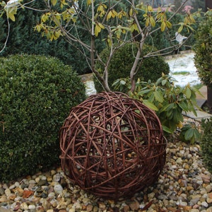 Könnte beinhalten: Eine große, braune, geflochtene Kugel aus Zweigen liegt auf einem Bett aus kleinen, bunten Steinen. Die Kugel ist von grünen Büschen und einem kleinen Baum umgeben.