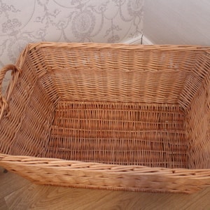 Pode incluir: Uma cesta de vime retangular grande com duas al&ccedil;as. A cesta &eacute; feita de vime natural tecido e tem uma cor castanho claro.