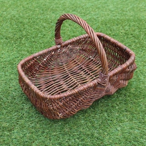 May include: A brown wicker basket with a handle. The basket is empty and sitting on a green grassy surface.