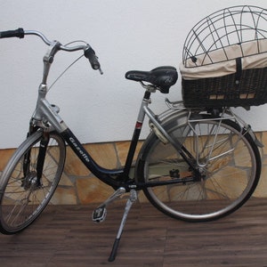Puede incluir: Bicicleta Gazelle plateada y negra con asiento negro y cesta trasera. La cesta tiene una jaula de metal y un forro de tela beige. La bicicleta tiene un caballete y guardabarros.