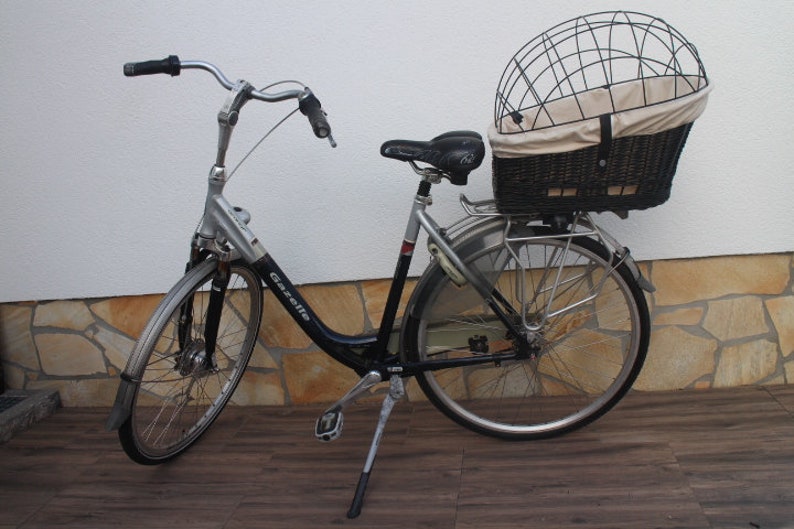 Puede incluir: Bicicleta Gazelle plateada y negra con asiento negro y cesta trasera. La cesta tiene una jaula de metal y un forro de tela beige. La bicicleta tiene un caballete y guardabarros.