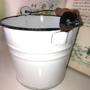 Könnte beinhalten: Weißer Emaille-Eimer mit schwarzem Rand und einem Drahtgriff mit Holzgriff. Der Eimer hat eine kleine, rechteckige Vertiefung an der Seite.