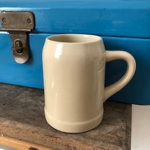 Könnte beinhalten: Ein cremefarbener Bierkrug aus Keramik mit Henkel. Der Krug hat eine kleine Inschrift auf der Seite, die "0,5l  Sachsen" lautet.