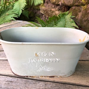 Könnte beinhalten: Ein weißer Metall-Pflanzkasten mit den Worten "Mon Jardin" in Prägung auf der Seite. Der Pflanzkasten ist oval geformt und hat ein rustikales, verwittertes Aussehen.