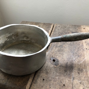 Könnte beinhalten: Ein vintage Aluminium-Topf mit einem Holzgriff. Der Topf hat ein abgenutztes, gealtertes Aussehen und steht auf einer Holzoberfläche.