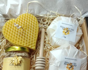 Könnte beinhalten: Eine Geschenkbox gefüllt mit Honig-basierten Bade- und Körperpflegeprodukten. Die Box enthält eine herzförmige Honigwabenkerze, ein Honigglas, einen hölzernen Honiglöffel und zwei weiße Seifenstücke, die in Papier eingewickelt sind. Die Seifenstücke sind mit "Honey Loofah Soap" und "Honey and Oats Hand Made Soap" beschriftet.