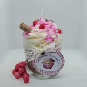 May include: A glass jar candle with a white and pink frosting design, topped with a chocolate bar, pink sprinkles, and a red raspberry. The label reads "Raspberry & Chocolate Cake" and "Handmade Soy Wax Candle".