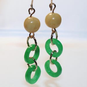 May include: A pair of dangle earrings with gold-colored hooks. Each earring features a light brown, round bead, connected to three bright green, circular jade stones. The earrings are suspended from a green stem.