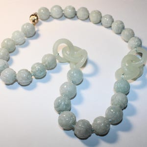 May include: A light green jade necklace with carved floral designs on the beads. The necklace features a gold clasp and a unique chain-link design. The beads are approximately 0.75 inches in diameter.