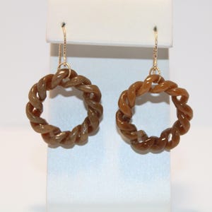 May include: A pair of earrings featuring a twisted rope design in a warm brown hue. The earrings are suspended from delicate gold-colored hooks. The earrings are displayed against a white background.