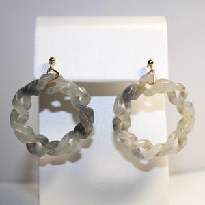 May include: A pair of gray and white hoop earrings with a twisted rope design. The earrings are made of a light-colored stone and have gold-toned clasps.