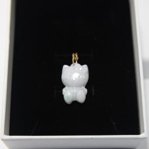 May include: A pale lavender and white carved jade pendant shaped like a cartoon cat character with a small bow on its head. The pendant features subtle green inclusions and is suspended from a thin gold-toned metal jump ring for attachment.