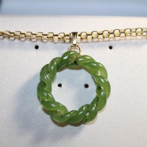 May include: A green jade pendant necklace with a gold chain. The pendant is a twisted, circular design. The necklace is displayed on a white surface.