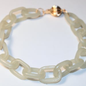May include: A light green chain link bracelet with a gold-colored clasp. The bracelet features large, rounded rectangular links. The clasp is a round, gold-colored magnetic closure. The bracelet is shown on a white background.