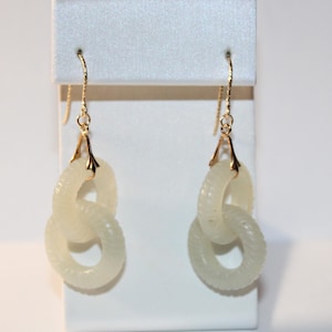 May include: A pair of gold-toned earrings featuring three interlocking, off-white, circular links. The links have a textured surface and are suspended from a gold-colored hook. The earrings are displayed on a white stand.