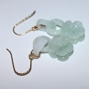 May include: A pair of gold-toned earrings with jade flower pendants. The earrings feature delicate gold hooks and are adorned with light green, carved jade flower charms. The earrings are displayed on a white surface.