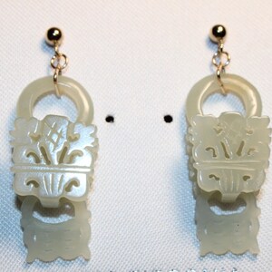 May include: A pair of jade earrings with gold-colored posts and connecting rings. The earrings feature a carved design with floral and geometric elements. The jade has a pale green hue, and the earrings are displayed against a white background.