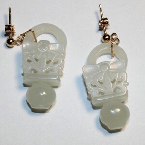 May include: A pair of white jade earrings with gold hooks. The earrings are shaped like a small vase with a carved design and a round bead at the bottom.