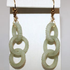 May include: A pair of jade earrings with a chain link design. Each earring features three interlocking, light green rings with a textured surface. The earrings have gold-colored hooks with decorative accents.