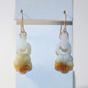 May include: A pair of jade earrings with a floral design. The earrings feature a gold chain and a white and yellow flower pendant. The earrings are displayed on a white stand.