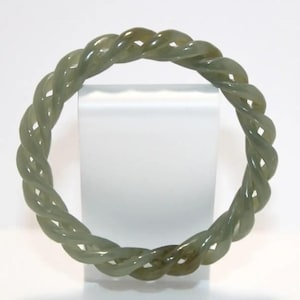 May include: A jade green bangle bracelet with a twisted rope design. The bracelet is a circular shape and appears to be made of a smooth, polished stone. The color is a muted green with some variations in tone.