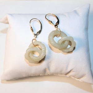 May include: A pair of dangle earrings featuring gold-tone leverback hooks attached to two interlocking pale green rings. Each earring consists of one smooth ring and one textured, ribbed ring, creating a layered, circular design with a soft finish.
