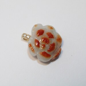 May include: A flower-shaped pendant with a white base and orange accents. The pendant has a gold-colored bail with small, clear stones. The design resembles a camellia or rose, suitable for jewelry.