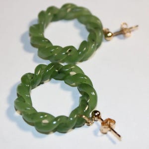 May include: A pair of green jade hoop earrings with a twisted design. The earrings are made of a light green jade stone and have a gold-tone post.