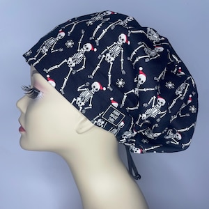 May include: Black scrub hat with a white skeleton pattern wearing a red Santa hat. The hat has a white snowflake pattern and the text "Ana Made This" is on the side.