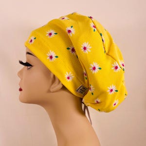 May include: A yellow scrub cap with a daisy pattern. The cap is made of a ribbed fabric and features white and pink daisies. The tag on the cap reads "AMA MADE THIS".