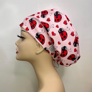 May include: A pink surgical scrub cap featuring a repeating pattern of red ladybugs and hearts. The ladybugs have black spots and antennae. The cap has a white drawstring and a small tag that says "ANIA MADE THIS".