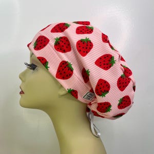 May include: A pink scrub cap with a repeating pattern of red strawberries with green stems. The cap has a ribbed texture and a white elastic band. The tag reads "ANA MADE THIS".