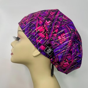May include: A vibrant purple and pink patterned scrub cap with gold accents. The cap features a geometric design and a small tag that reads "ANIA MADE THIS". The cap is designed to be worn in a medical setting.