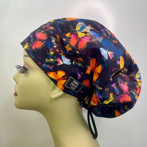 May include: A dark blue surgical scrub cap featuring a vibrant butterfly pattern in shades of orange, pink, yellow, and blue. The cap has a drawstring closure and a small tag that reads "ANIA MADE THIS."