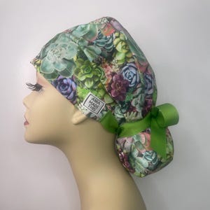 May include: A green, white, and purple surgical scrub hat with a succulent print. The hat has a green ribbon tied in a bow at the back. The hat has a white label that says "Ania Made This".