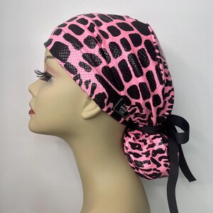 May include: A pink and black patterned scrub cap with a black ribbon tie. The cap features a snakeskin-like design and a small tag that reads "MADE THIS".